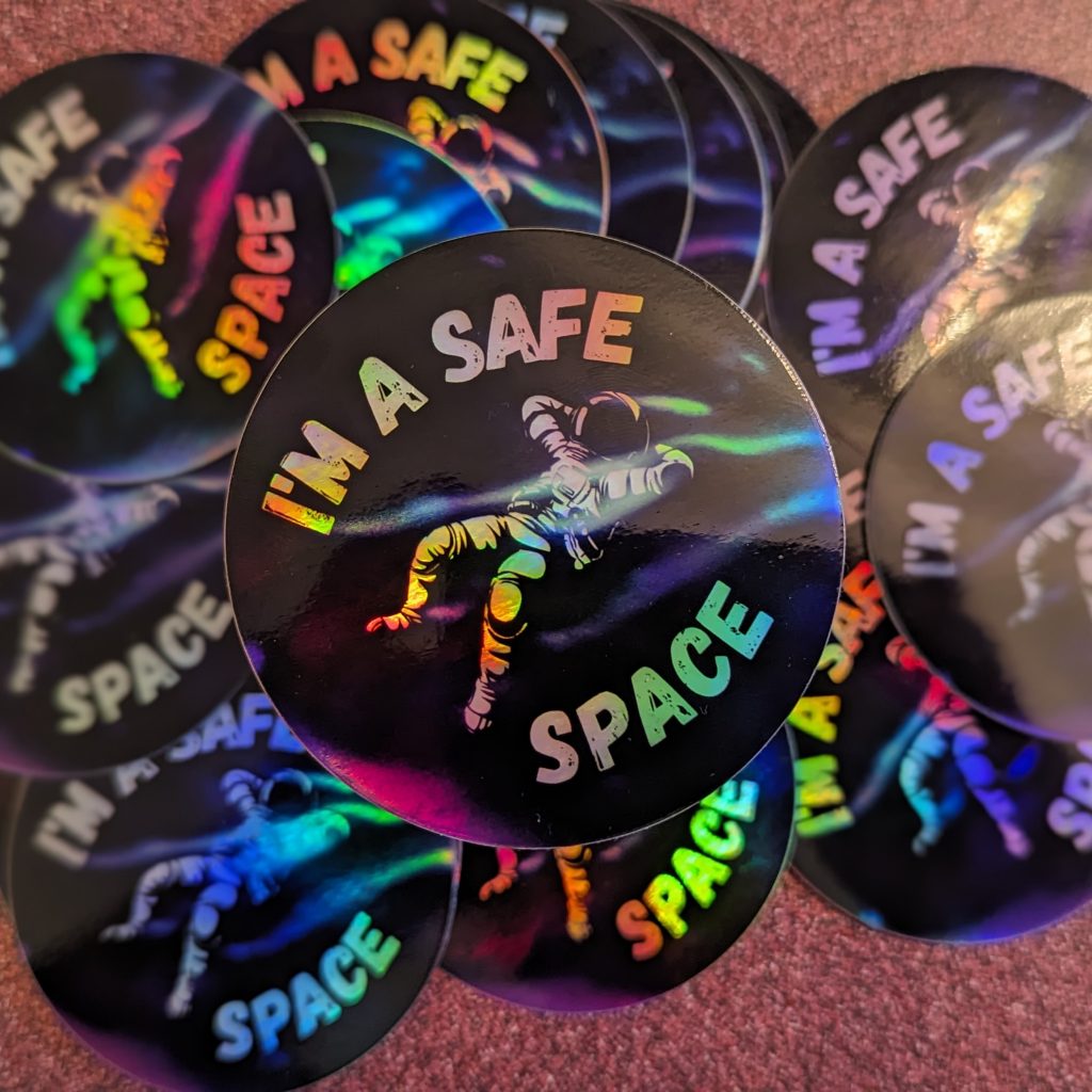 Product shot of a group of Holographic stickers. The stickers feature a relaxed astronaut and the phrase "I'm a Safe Space"